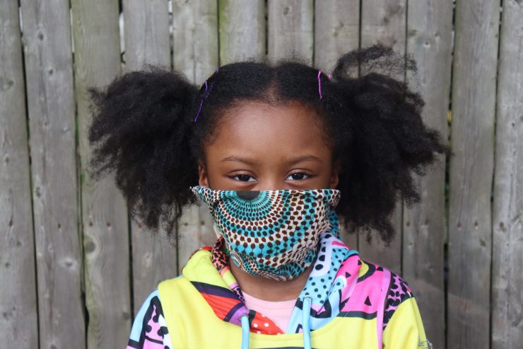 Cute African American Girl with pigtails wearing pattern fabric face mask outside with wooden fence background